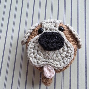 May include: A crocheted dog face with a tan, beige, and black color scheme. The dog has black button eyes, a black nose, and a pink tongue. The dog's face is round and has a textured appearance.
