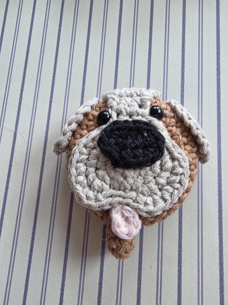 May include: A crocheted dog face with a tan, beige, and black color scheme. The dog has black button eyes, a black nose, and a pink tongue. The dog's face is round and has a textured appearance.
