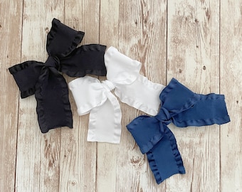 May include: Four large fabric bows in black, white, and navy blue, each with a ruffled edge. The bows are arranged on a light brown wooden surface, showcasing their contrasting colors and textures.