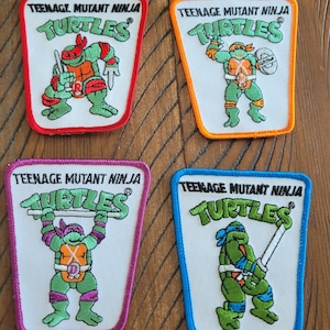 May include: Four embroidered patches featuring Teenage Mutant Ninja Turtles characters. Each patch has a white background with a different coloured border: red, orange, purple, and blue. The text "TEENAGE MUTANT NINJA TURTLES" is at the top of each patch.