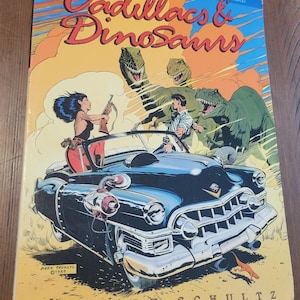 May include: A vintage comic book cover titled "Cadillacs & Dinosaurs" with a retro illustration. The artwork depicts a blue Cadillac with two people inside, pursued by dinosaurs. The cover has the text "Past and future meet in a world gone mad!" and "by Mark Schultz".