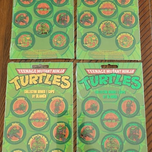 Può includere: Quattro confezioni di Teenage Mutant Ninja Turtles Collector Series 1 Caps di Slamco. Ogni confezione è verde e presenta la parola "Turtles" in un colore diverso. Ogni confezione contiene immagini circolari delle tartarughe.