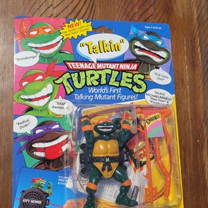 Op de afbeelding: Een vintage Teenage Mutant Ninja Turtles "Talkin'" actiefiguur, nog in de originele verpakking. De figuur is groen en oranje, met de verpakking met cartoonafbeeldingen van de schildpadden en de woorden "Kowabunga!" en "Radical, Dude!"