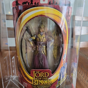 Könnte beinhalten: Eine Plastikvitrine mit einer Lord of the Rings Actionfigur. Die Figur zeigt einen Elfenkrieger in goldener Rüstung und einem roten Gewand. Die Figur hält ein Schwert und einen Schild. Die Verpackung zeigt das Lord of the Rings Logo und den Text "Die Gefährten".