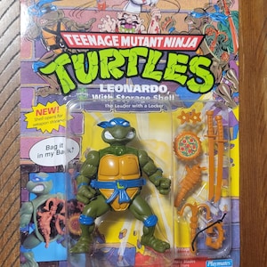 May include: Teenage Mutant Ninja Turtles Leonardo action figure with storage shell. The green turtle figure has a blue mask and comes with orange weapons and a pizza disc. The packaging includes the text "NEW!" and "Bag it in my Back!"