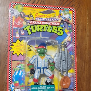 Könnte beinhalten: Teenage Mutant Ninja Turtles Actionfigur von Raphael in Baseball-Uniform. Die Figur befindet sich in einer Kunststoffverpackung mit den Aufschriften "Sewer Sports All-Stars" und "Grand Slammin' Raph".