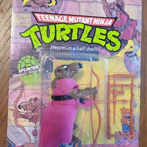 Könnte beinhalten: Teenage Mutant Ninja Turtles Splinter Actionfigur in Originalverpackung. Die Figur ist eine braune Ratte mit rosa Robe und schwarzem Gürtel, die einen Bogen hält. Die Verpackung zeigt die Aufschrift "Teenage Mutant Ninja Turtles" und "Heroes in a half shell!"