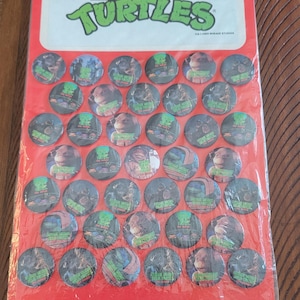 Könnte beinhalten: Eine Packung Teenage Mutant Ninja Turtles Buttons. Die Buttons sind rund und zeigen Bilder der Turtles. Die Verpackung ist rot mit den Worten "Teenage Mutant Ninja Turtles" in Grün und Weiß. Der Text "Button-Up" ist ebenfalls sichtbar.