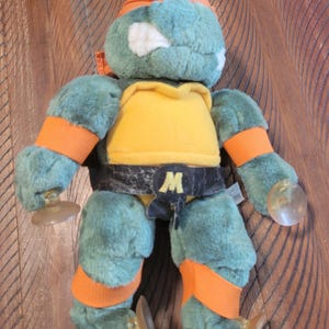 Op de afbeelding: Een pluche speelgoed van een Teenage Mutant Ninja Turtle, met een groen lichaam, oranje hoofdband en armbanden, en een gele borst. Het speelgoed heeft een zwarte riem met een witte "M" en doorzichtige zuignappen.