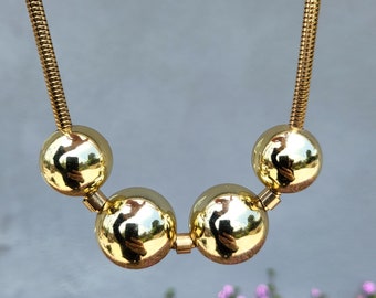 Puede incluir: Un collar de cadena de oro con cuatro cuentas grandes, redondas y doradas. Las cuentas son reflectantes y muestran una imagen borrosa del fotógrafo.