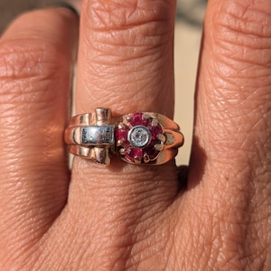 May include: A gold-toned ring with a central cluster of red gemstones surrounding a single, clear gemstone. The ring features a unique design with a silver-toned accent and is worn on a finger.