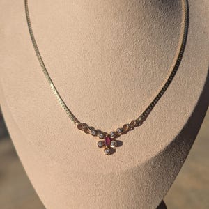May include: A gold necklace with a fine chain and a pendant. The pendant features a central ruby surrounded by small diamonds. The necklace is displayed on a beige bust. The pendant has a floral design. The necklace is a piece of jewellery.