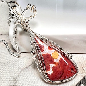 May include: A teardrop-shaped pendant featuring a polished red, white, and yellow patterned stone encased in intricate silver wire wrapping. The pendant hangs from a silver rope chain and includes decorative silver beads along the wire frame.
