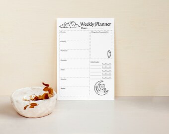 May include: A white paper weekly planner sheet featuring black ink illustrations of clouds, a crystal, and a cat sitting on a crescent moon. The layout includes sections for each day of the week, a gratitude list, and a habit tracker with circles.