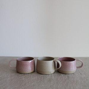 May include: Three ceramic mugs, one in a light beige color with speckles, and two in a light pink color. The mugs have a rounded shape and a handle on the side.