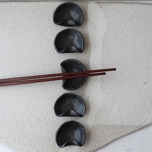 May include: Five black ceramic dipping bowls in a crescent shape, arranged vertically on a white surface. A pair of wooden chopsticks are resting on the third bowl from the top.