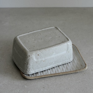 Könnte beinhalten: Ein rechteckiger, gesprenkelter, cremefarbener Keramik-Butterbehälter mit passendem Tablett. Der Deckel ist leicht abgerundet und sitzt auf einem Tablett mit strukturierter, gemusterter Oberfläche. Für die Küche.