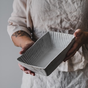 May include: A rectangular, light gray ceramic dish with a textured, ridged exterior. The handmade pottery piece is held by a person, showcasing its simple, yet elegant design. Ideal for serving or display.