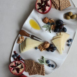 Könnte beinhalten: Eine weiße Servierplatte aus Keramik mit gesprenkelter Oberfläche und mehreren vertieften Fächern, angerichtet mit Käsescheiben, Walnüssen, Blaubeeren, Granatapfelhälften, grünen Oliven, Crackern und einer Käsegabel mit Holzgriff.