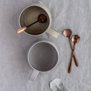 Könnte beinhalten: Zwei graue Keramikbecher mit Henkeln und ein roségoldener Teebeutel auf einem weißen Leinenhintergrund. Zwei roségoldene Löffel mit blumenförmigen Enden befinden sich ebenfalls auf dem Hintergrund.