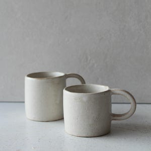 Könnte beinhalten: Zwei cremeweiße Keramikbecher mit abgerundeten Griffen. Die Becher haben ein schlichtes, minimalistisches Design und eine leicht strukturierte Oberfläche. Die Becher sind vor einem neutralen Hintergrund platziert, der ihre klaren Linien und Erdtöne hervorhebt.