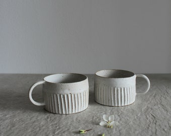 May include: Two white ceramic mugs with a textured, ribbed design. The mugs have a rounded shape and a handle on one side. The mugs are sitting on a light gray surface.