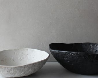 May include: Two ceramic bowls, one white and one black, with a textured surface. The bowls are both oval shaped and have a slightly raised rim.
