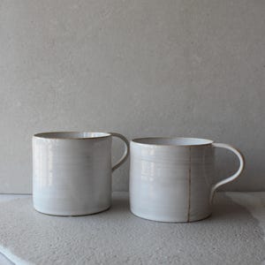 Peut inclure: Deux mugs en céramique blanche avec des anses arrondies. Les mugs présentent un design simple et minimaliste avec une surface légèrement texturée. Les bords des mugs ont une couleur brun doré subtile. Les mugs sont posés sur une surface gris clair.