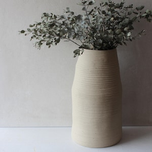 May include: A tall, cream ceramic vase with a textured, ribbed design. The vase is filled with leafy green branches, creating a natural and elegant display. The vase has a wide base and tapers towards the top.