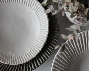 Può includere: Una pila di piatti in ceramica con un bordo a coste. Il piatto superiore è bianco, il piatto centrale è marrone scuro e il piatto inferiore è parzialmente visibile. I piatti sono su una superficie grigio chiaro, con una pianta sfocata sullo sfondo.