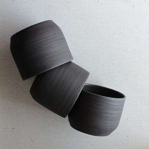 Peut inclure: Trois tasses en céramique gris foncé sont empilées sur une surface blanche. Les tasses ont une forme cylindrique avec un dessus légèrement incurvé et une surface texturée. Les tasses sont disposées en diagonale, l'une étant partiellement à l'intérieur de l'autre.