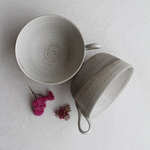 Puede incluir: Dos tazas de cerámica con un esmalte moteado. Las tazas son de color gris claro con un sutil patrón de espiral. Las tazas están sobre una superficie blanca.