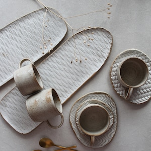 Peut inclure: Trois tasses en céramique blanche avec un glaçage moucheté et deux plateaux de service rectangulaires en céramique blanche avec un motif texturé.