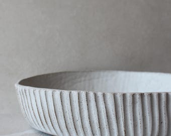 May include: A shallow, circular ceramic bowl with a matte off-white finish. The exterior features a distinct vertical ribbed texture with subtle dark speckles throughout the clay surface, creating a tactile and textured appearance on the vessel.