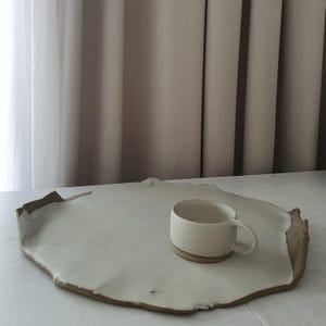 Può includere: Un vassoio da portata in ceramica bianco sporco fatto a mano con un bordo naturale e irregolare. Una tazza maculata abbinata con base e manico marrone è appoggiata sul vassoio. Lo sfondo presenta tende dai toni neutri.