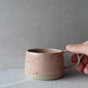 May include: A ceramic mug with a rounded shape and a side handle. The upper portion of the mug features a smooth, light pink glaze with small dark speckles, while the base is a contrasting, unglazed, textured beige material with dark flecks.