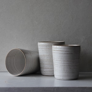 May include: Three handmade ceramic tumblers in a neutral color palette. The cylindrical cups have horizontal textured lines and a slightly speckled finish. One tumbler is lying on its side, while the other two stand upright.