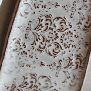 Può includere: Un vassoio rettangolare in ceramica con smalto bianco lucido su una base in argilla marrone scuro. La superficie è decorata con un motivo floreale in rilievo ripetuto di colore marrone scuro che rivela la trama dell'argilla sottostante.