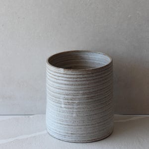 May include: A cylindrical ceramic vase with a textured, horizontally-lined surface. The vase is a light, off-white colour with subtle brown accents in the grooves. The open top reveals a smooth interior.