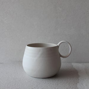 Puede incluir: Una taza de cerámica blanca con un cuerpo redondeado y un asa circular. La taza tiene un acabado mate y un diseño simple y minimalista. La taza es adecuada para café, té u otras bebidas.