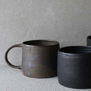 Könnte beinhalten: Nahaufnahme von drei Keramikbechern in Schwarz- und Brauntönen. Der Becher links hat einen Henkel und eine strukturierte Oberfläche. Die beiden anderen Becher sind henkellos und haben eine glatte, matte Oberfläche. Die Becher stehen auf einer hellgrauen Oberfläche.