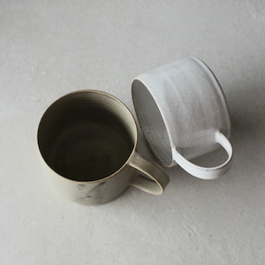 Op de afbeelding: Twee keramische mokken, een in een lichtgrijze glazuur en de andere in een donkergroene glazuur. Beide mokken hebben een afgeronde vorm en een handvat.