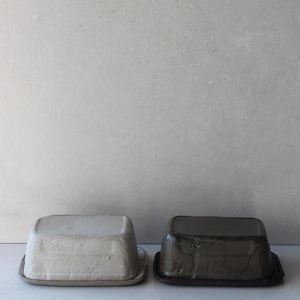 Peut inclure: Deux beurriers en céramique, l'un de couleur gris clair et l'autre de couleur gris foncé. Les deux beurriers ont une surface texturée et une forme rectangulaire.