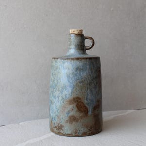 Może przedstawiać: Ceramiczna butelka z korkiem i małym uchwytem. Butelka ma niebiesko-brązową glazurę o teksturowanym wyglądzie. Design przypomina zabytkowy dzbanek, odpowiedni do celów dekoracyjnych.