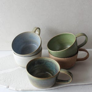 Può includere: Una collezione di tazze in ceramica fatte a mano in vari colori. Le tazze hanno una forma arrotondata con un manico e sono impilate. I colori includono blu, verde e marrone. Adatte per caffè, tè o altre bevande.