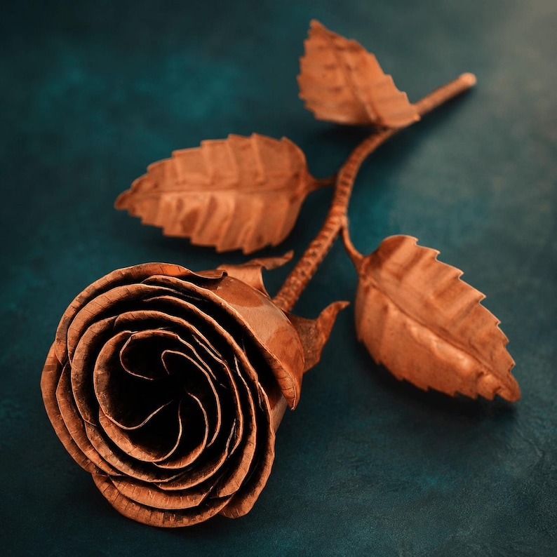 May include: A handcrafted copper rose sculpture featuring a tightly coiled bloom with layered petals and a slender stem. The metal flower includes three distinct serrated leaves attached to the textured stalk, showcasing a warm metallic finish.