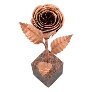 Könnte beinhalten: Eine kupferfarbene Metallrosen-Skulptur mit detaillierten Blütenblättern und Blättern. Die Rose steht auf einem strukturierten, quadratischen Sockel. Der Stiel und die Blätter weisen komplizierte Aderdetails auf, die ein realistisches Aussehen erzeugen. Die Skulptur ist ein dekorativer Artikel.