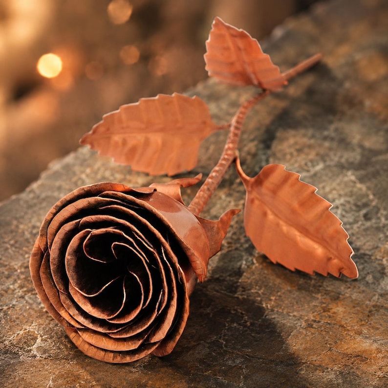 May include: A handcrafted copper rose sculpture featuring a detailed bloom with layered petals, a textured stem, and three distinct serrated leaves. The metallic surface displays a warm, reddish-brown finish with subtle variations in light and shadow.