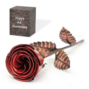 May include: A metal rose with a deep red bloom and bronze stem and leaves. A small, textured box with the words "Happy 8th Anniversary" is also present. The rose is a romantic gift, ideal for an anniversary.