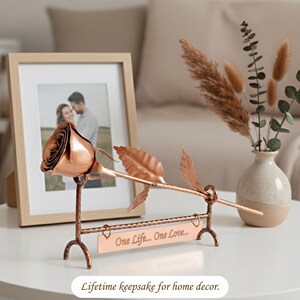 May include: A copper-coloured metal rose sculpture resting on a matching stand with a rectangular plaque engraved with the text One Life... One Love... The rose features a single bloom and two leaves on a stem, supported by a textured metal frame.